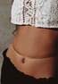 A close-up of a person's midriff wearing a white lace crop top, black bottoms, and the AHIMSA CHOKER gold plated chain waist accessory against a neutral background.