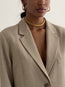 A woman is shown from the shoulders up against a plain background, wearing a beige blazer, gold hoop earrings, and the AHIMSA CHOKER—a layered choker featuring a gold plated chain.