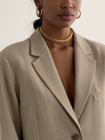 A woman is shown from the shoulders up against a plain background, wearing a beige blazer, gold hoop earrings, and the AHIMSA CHOKER—a layered choker featuring a gold plated chain.